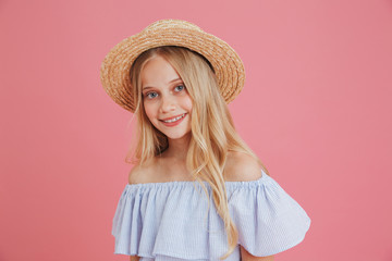 Image of lovely blond summer girl 8-10 wearing blue dress and straw hat smiling at camera with joyful glance, isolated over pink background