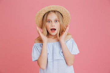 Image closeup of excited european girl 8-10 wearing blue summer dress and straw hat raising hands at face in surprise, isolated over pink background