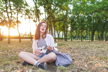 Portrait of a beautiful European girl sitting in a park playing ukulele and singing. Sympathetic girl musician sits on herbs in a park with Hawaiian guitar. Musical concept