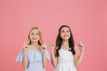 Image of two caucasian girls 8-10 years old wearing dresses looking upward with smile and pointing fingers at copyspace, isolated over pink background