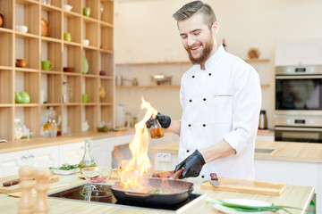 Portrait of smiling  professional chef working in modern restaurant kitchen standing at wooden table and cooking flambe dish, copy space