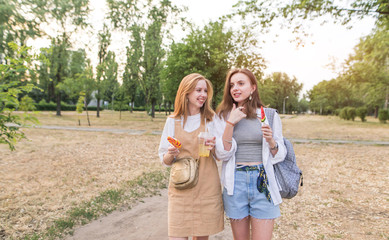 Fototapeta premium Two stylish girlfriends in light clothing with lemonade and lollipops stroll through the summer evening park. Two stylish attractive girls walking around the park and smiling.