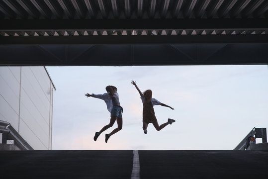 Silhouette of two young girls who jump on. Silhouette of two friends in a jump on the background of the sky near modern architecture. - Powered by Adobe