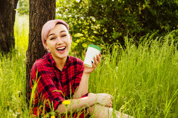 Young beautiful smiling woman sitting in green grass and holding paper cup with coffee on a tree and bush background