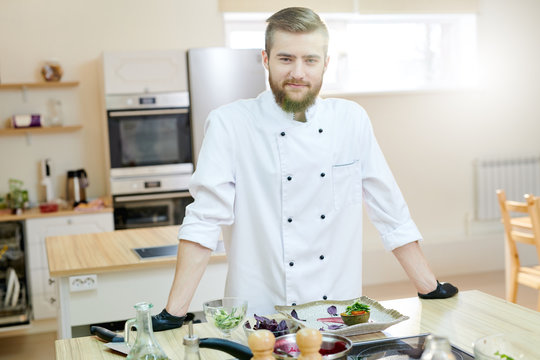 Portrait of handsome bearded chef posing in kitchen standing at wooden table and looking at camera, copy space
