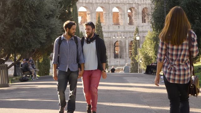Young happy gay couple tourists walk in park road with trees colosseum in background in rome at sunset holding hands gelousy when beautiful attractive girl walks by glancing lovely slow motion colle