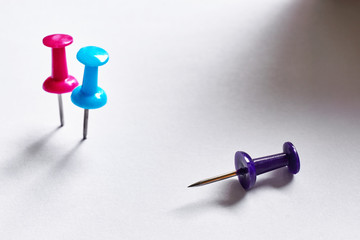 Close-up of two standing blue and red colored push pins and one overturned purple pin on a white background with dramatic shadow 