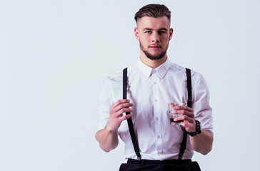 Confident, stylish man in white shirt and suspenders looking to camera and holding glass of whiskey in hand while sitting against gray background. Elegant man drinking alcohol in studio. Men's fashion