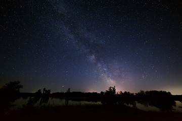 Space with stars in the night sky. The landscape with the river and trees is photographed on a long exposure.