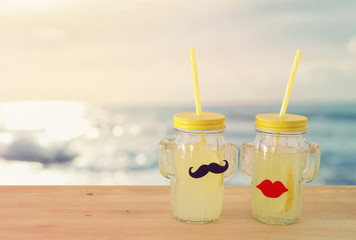 Image of fresh lemonade drink in cute cactus shape glasses wearing mustache and lips, over wooden table. Tropical summer romantic vacation concept.