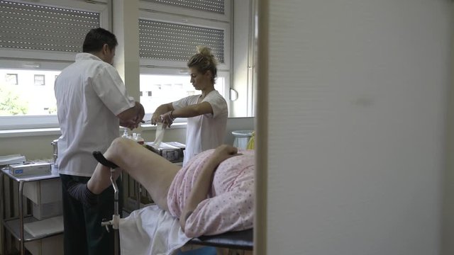 Male gynecologist prepares for gynecological examination of woman, nurse puts protective glove on doctor hand, female patient lying on medical review in ambulance room at gynecology ward, health care.