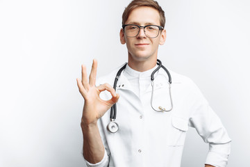 Portrait of a young doctor, on a white background, which shows the gesture Okey, trainee in the Studio, with a stethoscope on the neck, for advertising, text insertion