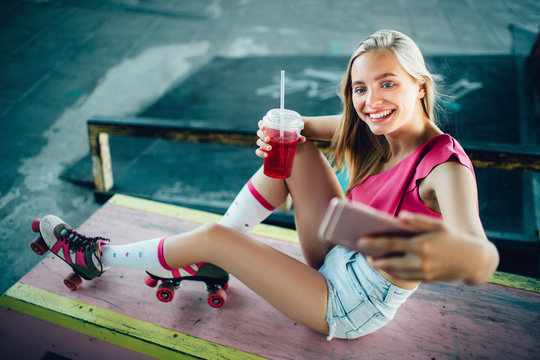 Horizontal picture of pretty blonde girl sitting on pink ground and taking selfie. She is smiling on camera. There is a cup of red drink in her right hand. She is happy.