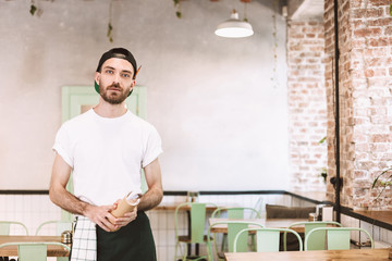 Young waiter in black cap,white t-shirt and apron standing with notepad in hands and dreamily...
