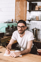Young smiling man in eyeglasses and cap standing behind bar counter with pencil and notepad and...
