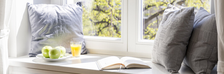 Real photo of windowsill with three grey pillows, open book, green apples on plate and orange juice...