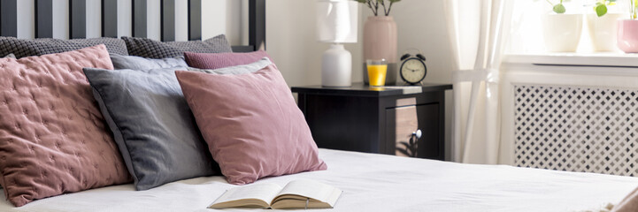 Open book placed on double bed with pastel pillows standing in the real photo of bright bedroom interior with clock, white lamp and juice in glass on dark bedside table