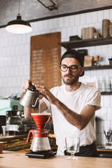 Young barista in eyeglasses and cap standing at bar counter and preparing pour over coffee while...