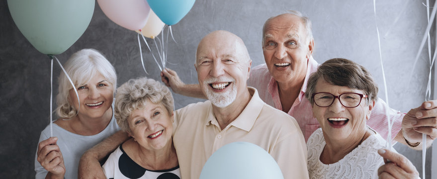 A group of happy, senior friends holding colorful balloons while posing at a party and celebrating birthday together.