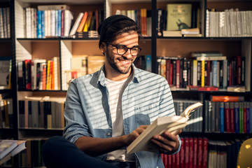 Young handsome male student is sitting in a library near the bookshelf and is enjoying his book.