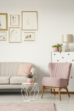 Book and plant on a diamond shape side table, beige sofa and comfy armchair in an elegant, white living room interior with framed posters gallery