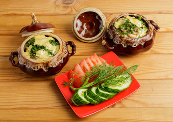 Potatoes are baked in a pot of meat and cheese and a plate of cucumbers and tomatoes on a wooden table.