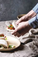 Man's hands hold asian sandwich steamed gua bao buns with pork belly, greens and vegetables served in ceramic plate on table with linen tablecloth. Asian style fast food dinner.