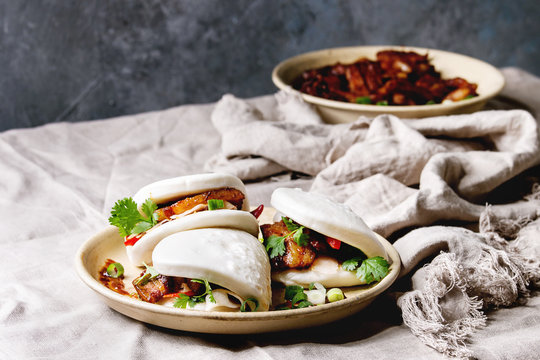 Asian sandwich steamed gua bao buns with pork belly, greens and vegetables served in ceramic plate on table with linen tablecloth. Asian style fast food dinner.
