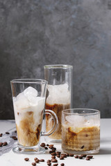 Iced coffee cocktail or frappe with ice cubes and cream served in three different glasses with coffee beans around on white marble table with grey concrete wall at background.