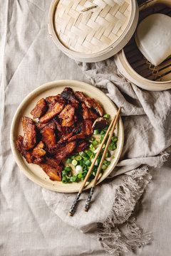 Chinese traditional dish Cantonese BBQ Pork Belly with spring onion served in ceramic plate with chopsticks, bamboo steamer and gua bao bus over linen cloth. Flat lay, space. Asian style dinner.