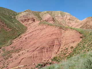 The mountain with red scree slopes covered by green grass. The sky is clear and blue. It is the sunny summer day.