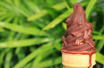 Melting Chocolate Dipped Soft Serve Ice Cream Cone with Blurred Green Bush in Background 