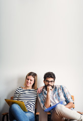 Handsome man and cheerful beautiful woman are waiting for their job interview while sitting on chairs and holding folders on their laps.