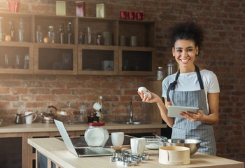 Happy black woman baking with recipe in tablet