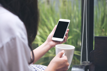 Mockup image of a woman holding white smart phone with blank black desktop screen while drinking coffee with blur green nature background