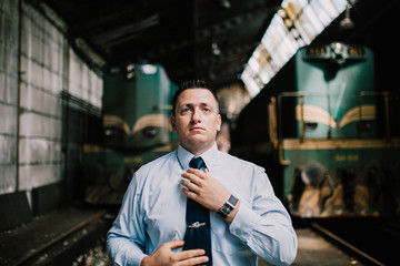 Young railroad engineer tie a tie in old locomotive depot with two locomotives behind him