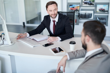Portrait of bearded handsome sales manager talking to client while sitting at desk in car showroom