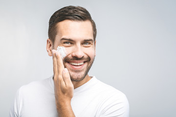 Skin care. Handsome young shirtless man applying cream at his face and looking at camera with smile...