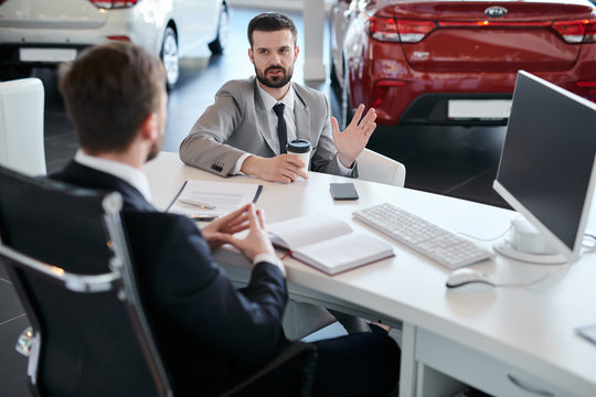 Portrait Of Mature Bearded Businessman Talking To Sales Manager Sitting Across Desk  In Car Dealership Showroom, Copy Space