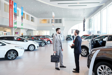 Full length portrait of handsome car salesman talking to customer in luxury showroom, copy space