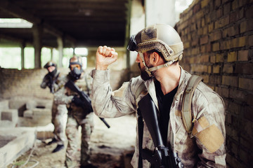 Strong guy is standing in opened big room with his comrades and looking to them. He is shwoing fist. Other guys are looking at him and holding black rifles.