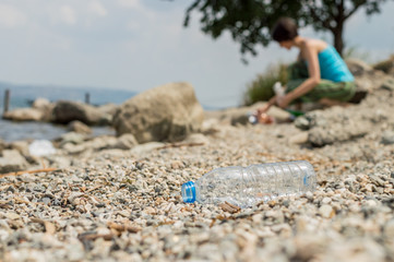 Plastic bottle dumped on the coastal strip with blurry woman picking up the garbage on the background.