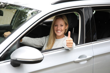 Hübsche blonde Frau am Steuer eines Autos lächelt und zeigt Daumen hoch