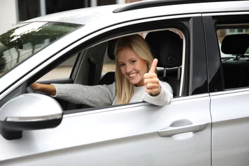 Fotobehang Hübsche blonde Frau sitzt am Lenkrad eines Autos und zeigt lachend Daumen hoch © Joerch