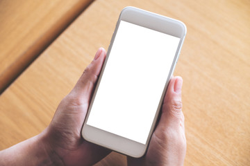 Mockup image of hands holding white mobile phone with blank desktop screen on wooden table in cafe
