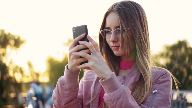 Pretty girl in glasses holding smartphone in hands. Young Caucasian attractive woman in pink jacket taking pics of herself on background of beautiful park. Springtime.