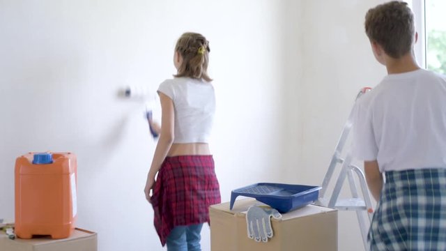 Teen boy and girl makes repairs in new flat. Sister and brother painting wall in apartment room. Children paints the wall, using a rollers. Young friends laughing and having fun. 