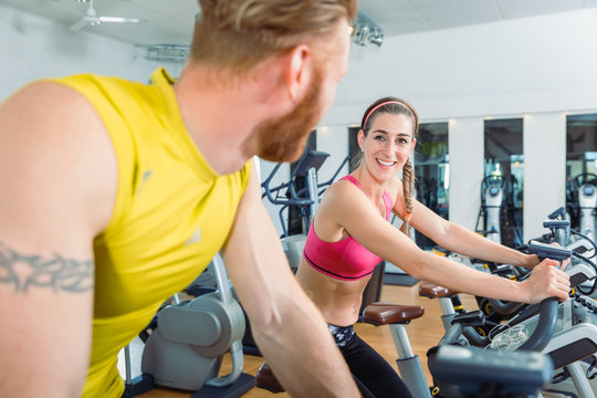 Beautiful fit woman smiling at the handsome man next to her while cycling on stationary fitness bicycles during cardio training at the gym