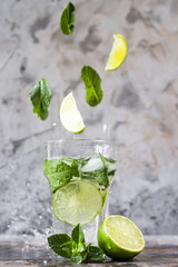 Leaves of mint and lime fly into a glass with mojito with ice on a gray background. Levitation flying magic items. Summer drink. With a spray of water
