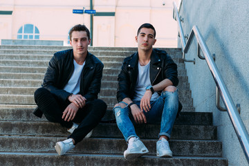 two guys in black jackets, white T-shirts, jeans and white shoes are sitting on the steps. on the hands of a watch. socializing, meeting friends.